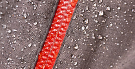A close-up photo of a piece of a fabric with many droplets of water on it. The fabric is brown with a red stripe.
