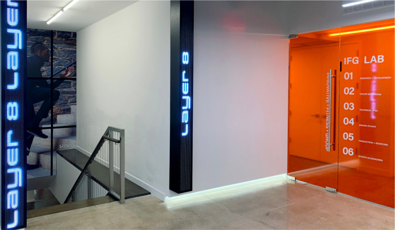 A photo of the Adjmi Design Studio. On the left is a stairwell leading to the Layer 8 division, with vertical marquees on each side of the stairs. On the right is an orange glass door which leads to the IFG Lab.