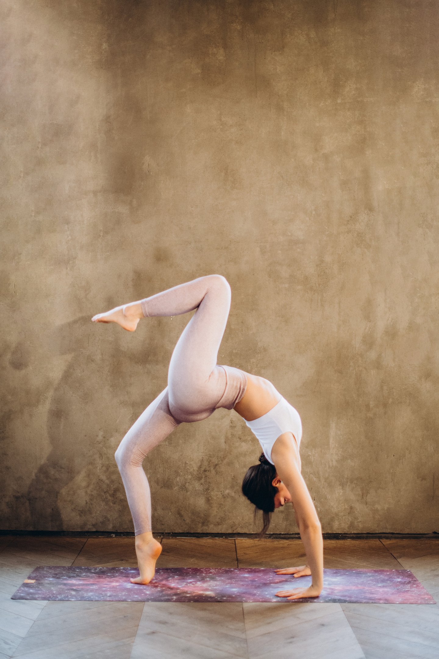 A side profile photo of a yoga practitioner performing a one-legged bridge pose on a yoga mat in front of a beige, rock-textured wall. She is wearing light pink yoga pants and a white tank top.