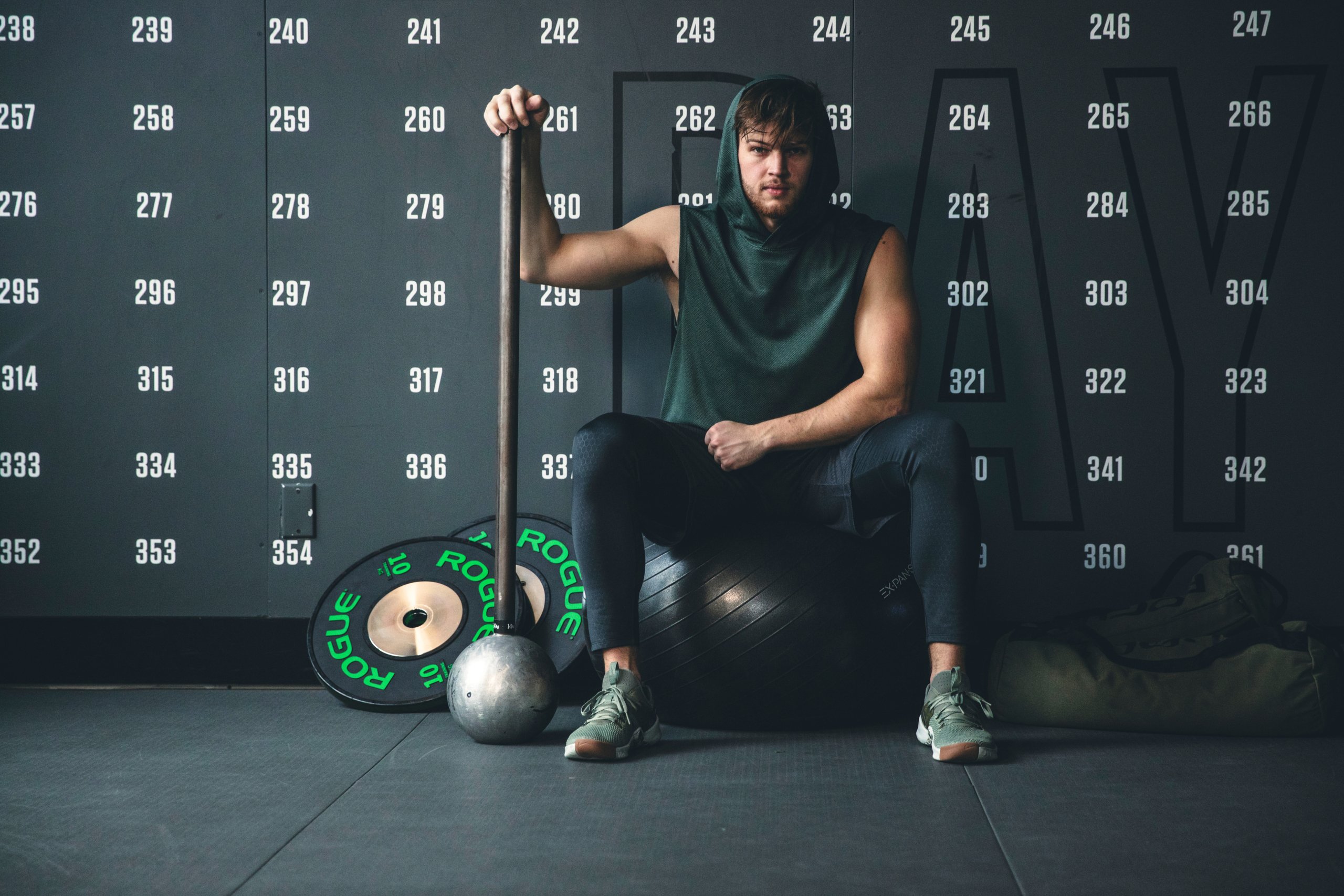 A photo of an athlete sitting on an exercise ball. He is facing the camera and grasping a large mace-bell with his right hand. He is dressed in a dark green sleeveless hoodie, black training shorts with black compression leggings underneath, and green sneakers. A couple of large weight plates are stacked behind the mace-bell.